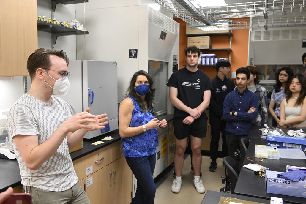 A man with brown hair and glasses is standing in front of scientific equipment, speaking to a group of students in a laboratory. His hands are held in front of him as if he is making very important gestures while speaking.