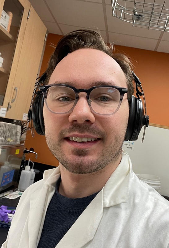 A selfie of a man with glasses wearing a labcoat and wireless headphones in a laboratory.