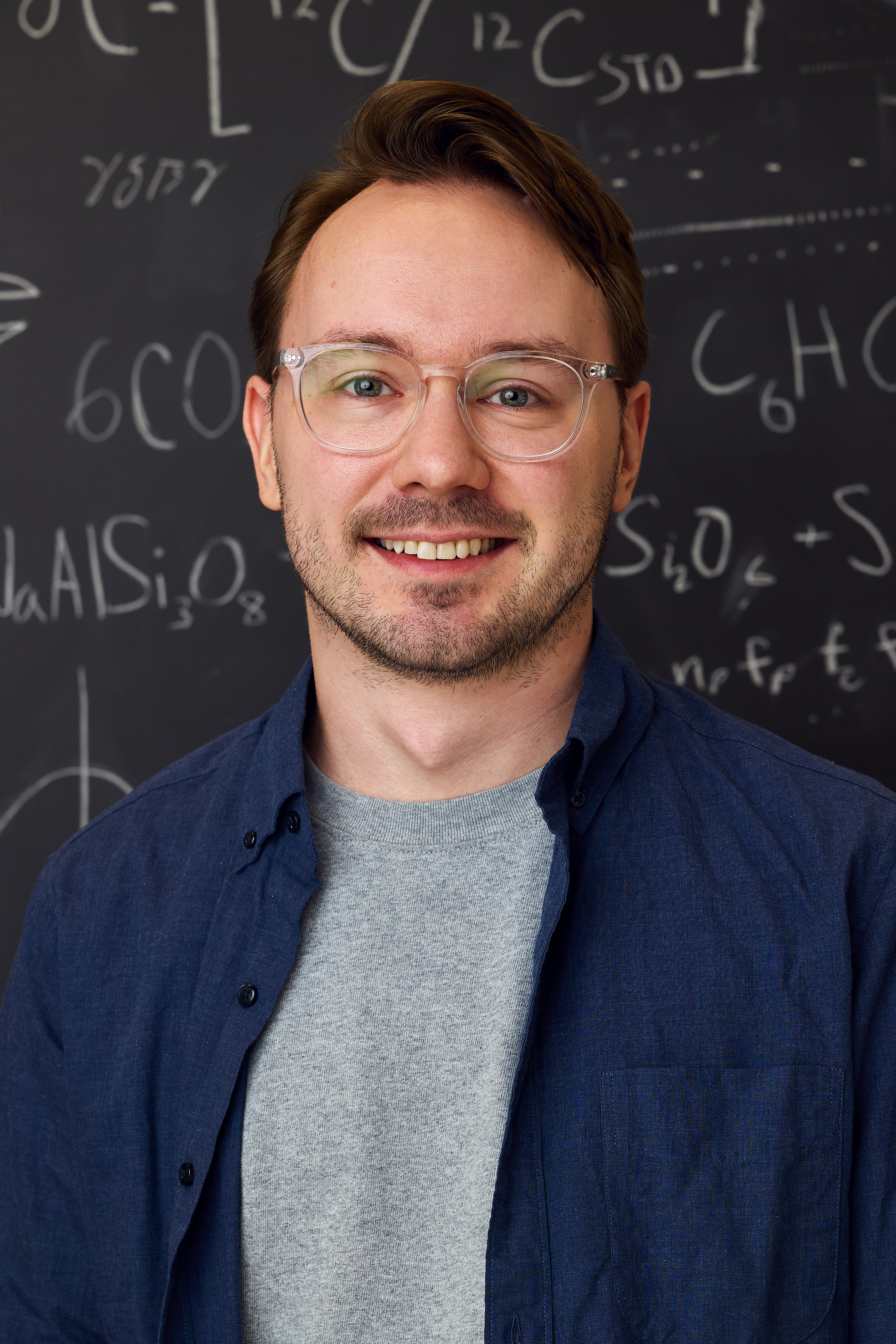 Photo of a man with brown hair and glasses standing in front of a blackboard with equations written on it.